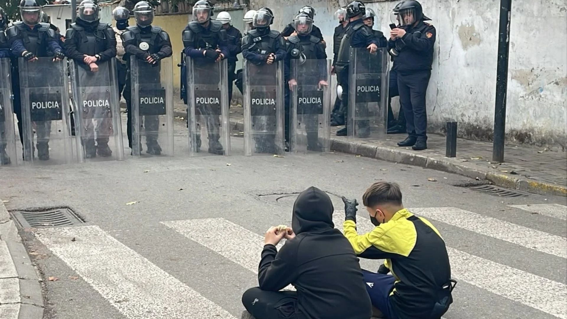 Protesta para Ministrisë së Arsimit  Dy protestues të vegjël të maskuar ulen këmbëkryq para efektivëve  një tjetër valëvit flamurin shqiptar pas tyre